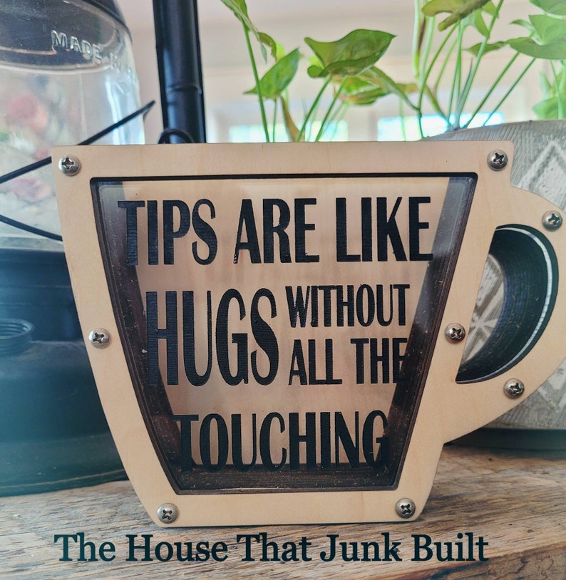 May include: A wooden sign shaped like a coffee mug with the text "Tips are like hugs without all the touching". The sign is painted in a light brown colour and has a dark brown border. The text is in black.