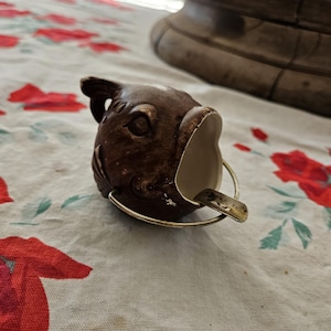 May include: A brown ceramic fish-shaped creamer or pitcher with a metallic gold-toned finish on the raised details. The vessel features a wide open mouth serving as a spout and a thin, curved metallic handle attached to the sides of the fish body.