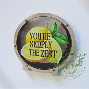 May include: Round wooden sign with a clear front featuring a lemon illustration and the text "YOU'RE SIMPLY THE ZEST." The sign includes a green leaf and is mounted with silver screws. The sign has a light wood frame.