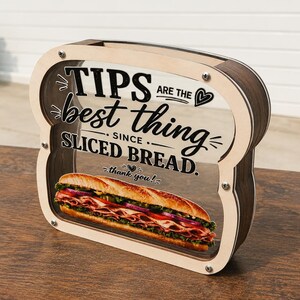 May include: A bread-shaped wooden tip jar with a clear front panel featuring the black text "TIPS ARE THE best thing SINCE SLICED BREAD. thank you!" and a graphic of a sub sandwich. The frame is light wood with dark wood sides and metal screws.