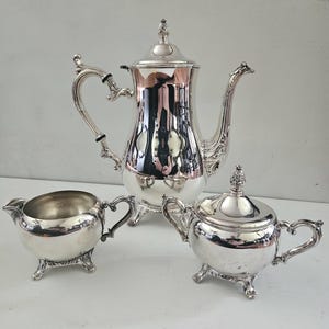 May include: A polished silver tea set consisting of a tall teapot with a lid, a creamer, and a lidded sugar bowl. Each piece features ornate, curved handles, decorative finials on the lids, and stands on three small, detailed feet.