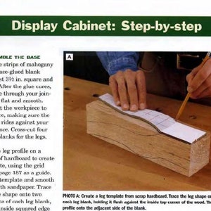 May include: A step-by-step guide for assembling a display cabinet. The image shows a person tracing a leg shape onto a piece of wood using a template. The text in the image reads "Assemble the Base" and "Draw the leg profile".