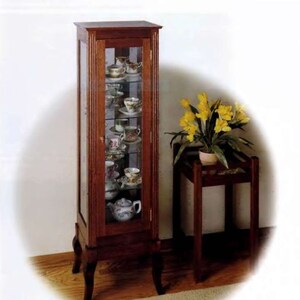 May include: A tall, wooden display cabinet with glass doors and shelves. The cabinet is filled with teacups and saucers, and a teapot. The cabinet has a dark wood finish and sits on four legs.