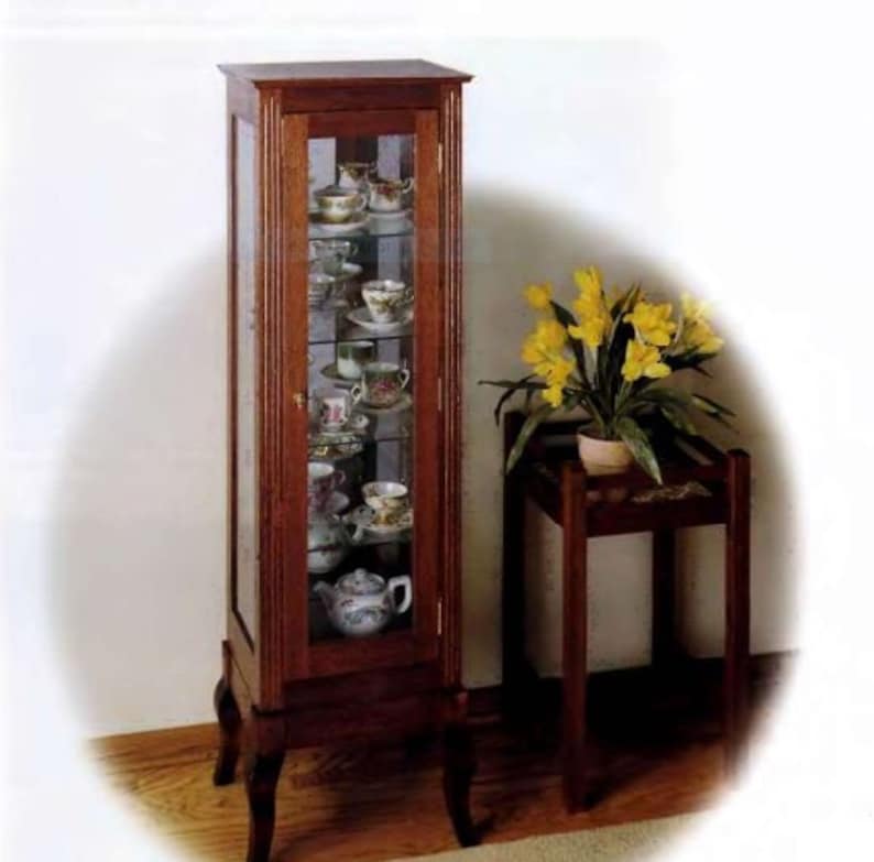 May include: A tall, wooden display cabinet with glass doors and shelves. The cabinet is filled with teacups and saucers, and a teapot. The cabinet has a dark wood finish and sits on four legs.