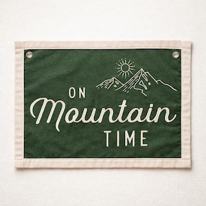 May include: A rectangular green banner with the words "ON Mountain TIME" in white script. The banner features a white outline of mountains and a sun. The banner has a cream-colored border and metal grommets.