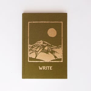 May include: An olive green hardcover notebook featuring a gold foil stamped illustration of a mountain range under a circular sun. The word WRITE is printed in gold capital letters below the rectangular mountain graphic on the front cover.