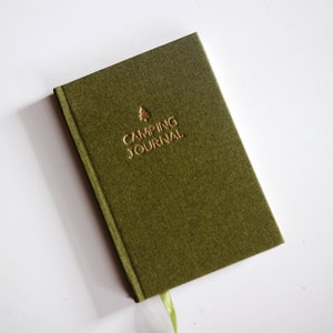 May include: Olive green camping journal with gold lettering and a tree icon. The words "CAMPING JOURNAL" are embossed on the cover. A green ribbon bookmark is visible. The journal is on a white surface.