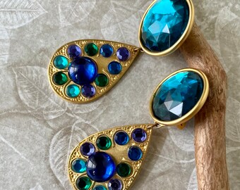 May include: A pair of gold tone dangle earrings with blue and green rhinestones. The earrings feature a large blue rhinestone at the bottom and a smaller blue rhinestone at the top.