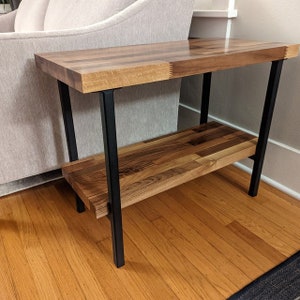Puede incluir: Una mesa auxiliar de madera con estructura de metal negro. La mesa tiene dos estantes y est&aacute; hecha de madera clara y oscura.