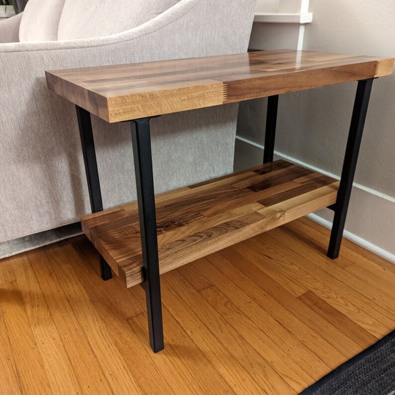 Puede incluir: Una mesa auxiliar de madera con estructura de metal negro. La mesa tiene dos estantes y est&aacute; hecha de madera clara y oscura.