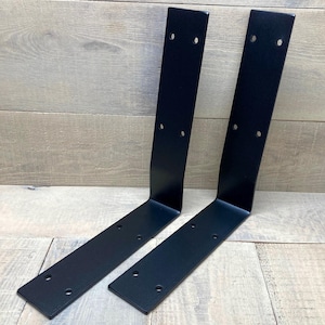 May include: Two L-shaped metal shelf brackets with a matte black finish. Each bracket features a vertical arm and a horizontal arm, both containing multiple pre-drilled circular holes for mounting purposes. The brackets are positioned side by side.