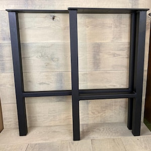 May include: Two black metal table legs with a rectangular frame design. The legs are made of square tubing and have a horizontal support bar. The legs are designed to be attached to a tabletop.