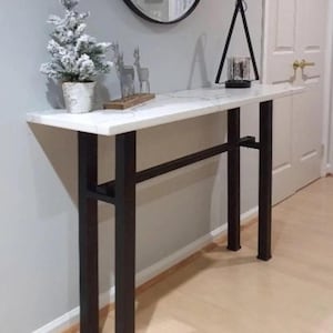 May include: A modern console table with a white marble-look top and black metal legs. The table has a rectangular shape and a connecting bar for stability. A small decorative Christmas tree and a black geometric candle holder are on the table.