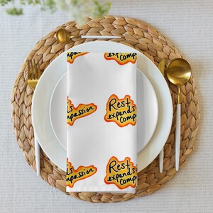 Könnte beinhalten: Weißes Stoffserviette mit den Sätzen "Rest expands comp" und "Is passion" in gelben und orangen Buchstaben. Die Serviette liegt gefaltet auf einem weißen Teller, mit goldenem Besteck und einem gewebten Platzset.