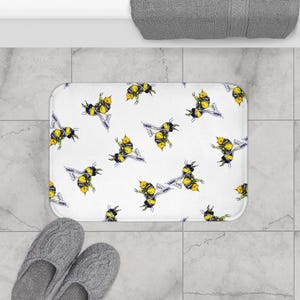 May include: White bath mat with a repeating pattern of yellow and black bumblebees. The bees are drawn in a watercolor style.