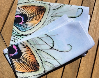 May include: A folded fabric napkin featuring a printed peacock feather pattern. The design displays a central eye motif with shades of orange, black, and pink, surrounded by light blue and green wispy lines on a white background.