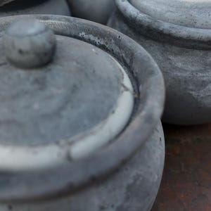 May include: Close-up of several gray ceramic pots with lids. The pots have a textured, weathered appearance, with a small knob on top of each lid. The pots are arranged closely together, suggesting a set or collection.