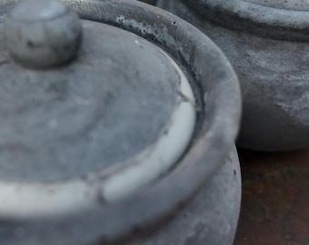 May include: Close-up of several gray ceramic pots with lids. The pots have a textured, weathered appearance, with a small knob on top of each lid. The pots are arranged closely together, suggesting a set or collection.
