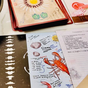 May include: A stack of recipe cards with a floral design and handwritten notes. One card features a lobster illustration and the word "Shrimp." Other cards show recipes and cooking times. The cards have red edges.
