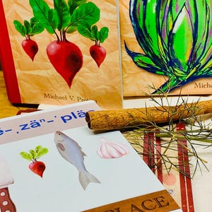 May include: A stack of illustrated cards and two books titled "The Chef Oracle" are displayed. The cards feature images of food items like a radish, fish, and garlic. A cinnamon stick and sprigs of dill are also present.