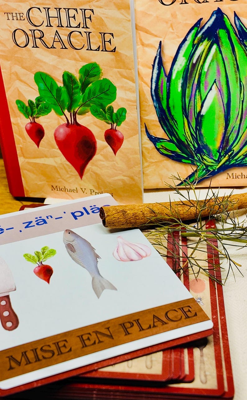 May include: A stack of illustrated cards and two books titled "The Chef Oracle" are displayed. The cards feature images of food items like a radish, fish, and garlic. A cinnamon stick and sprigs of dill are also present.