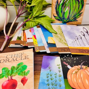 May include: A collection of "The Chef Oracle" cards and a book, featuring colorful illustrations of vegetables, herbs, and kitchen tools. Fresh basil with purple stems and green leaves is in the foreground. The cards have text like "ROS" and "SPO".