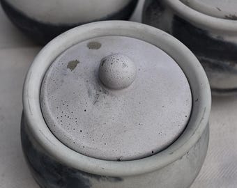May include: Three small, round, lidded containers in shades of gray. The containers have a textured, concrete-like appearance with a dark gray band around the base. Each lid has a small, round knob.
