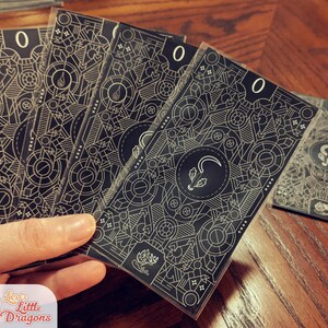 May include: Five black and gold playing cards with a geometric pattern and the number "0" printed on each card. The cards are held in a hand.