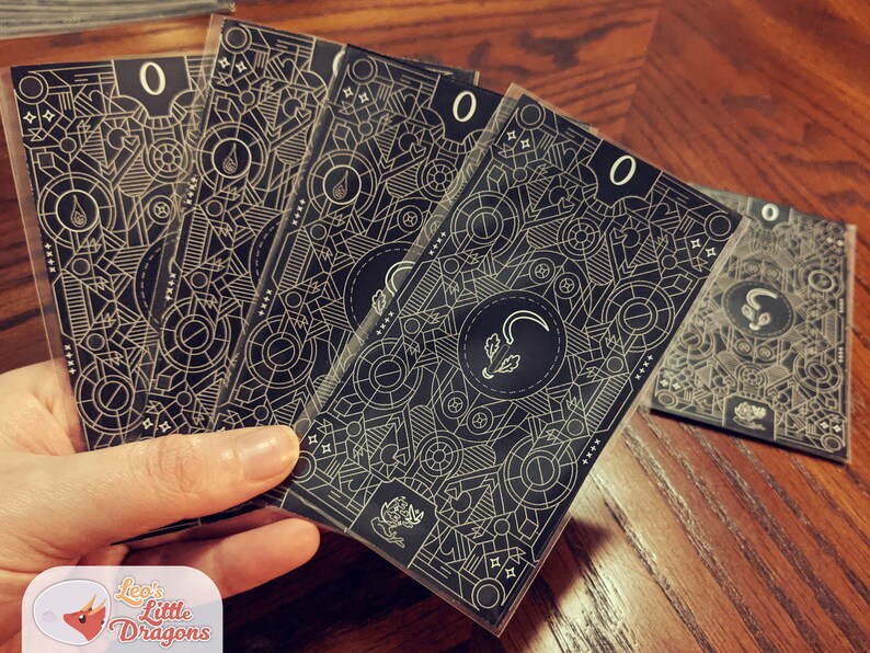 May include: Five black and gold playing cards with a geometric pattern and the number "0" printed on each card. The cards are held in a hand.