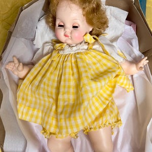 May include: A vintage doll with blonde, curly hair and closed eyes, wearing a yellow and white gingham dress with a white collar and cuffs. The doll is lying in a cardboard box, with its arms outstretched.