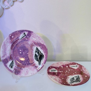 May include: Two decorative plates with a seashell theme. The round plate and the oval plate have a marbled purple and white background with black seashell illustrations. The plates are displayed on clear stands.