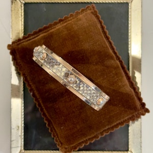 May include: A rectangular gold-toned barrette with a geometric pattern and clear gemstones, resting on a brown velvet pillow inside a gold-framed mirror. The barrette has a decorative design.