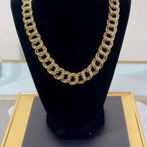 May include: A gold-colored chain necklace with a double-link design is displayed on a black velvet bust. The necklace is made of a shiny, metallic material. The bust is on a black rectangular base, set against a neutral background.