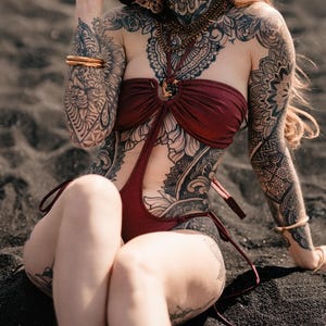 May include: A woman wearing a burgundy swimsuit and a black sun hat sits on black sand. Intricate tattoos cover her body, arms, and neck. She wears gold bracelets and a necklace. The swimsuit has a halter top design.