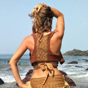 May include: A brown and tan crop top with a decorative back panel and a matching wrap skirt. The outfit is worn on a beach with the ocean in the background. The top has a bow in the back.