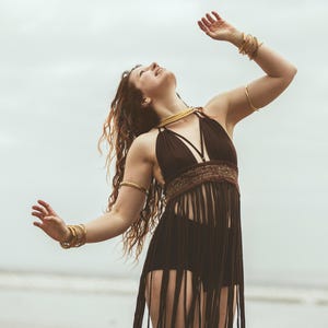 May include: A dark brown halter top featuring a textured midsection and long fringe detailing that extends to the thighs, paired with matching dark brown high-waisted bottoms, accented by multiple gold-toned arm cuffs and a gold-toned choker necklace.