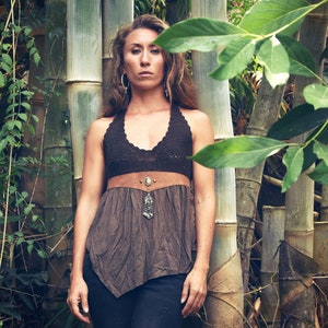 May include: A woman wearing a brown crocheted halter top with a brown leather belt and a silver pendant necklace. She is standing in front of a bamboo grove.