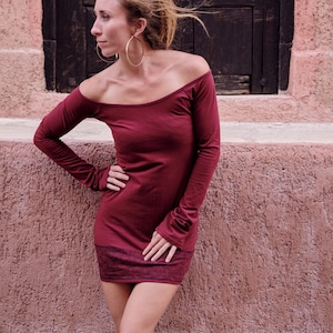May include: A burgundy off-the-shoulder long-sleeve dress with a fitted silhouette. The dress features a scoop neckline and a textured hemline. Large gold hoop earrings are worn. The background includes a weathered wooden door and a textured pink wall.
