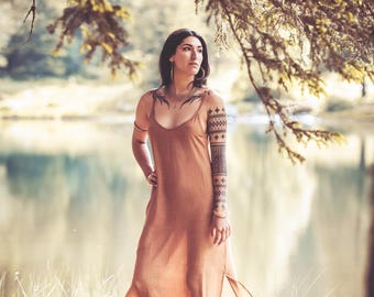 May include: A woman wearing a long, light brown dress stands on a rock by a lake. She has a geometric tattoo on her arm and a necklace with a pendant.