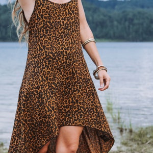Modèle portant une robe à bretelles mi-longue, coupe asymétrique, imprimé léopard marron, vue de face trois quarts. Décor bord de lac, herbe et sable, arbres au loin.