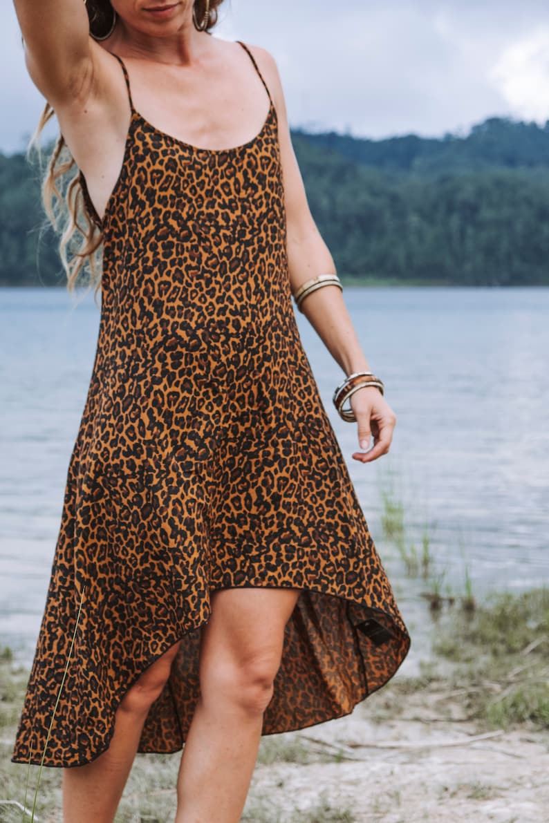 Modèle portant une robe à bretelles mi-longue, coupe asymétrique, imprimé léopard marron, vue de face trois quarts. Décor bord de lac, herbe et sable, arbres au loin.
