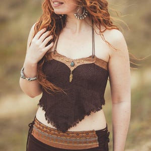 May include: A brown halter top with a fringed hem and a decorative neckline. The top features a brown lace design with a leather-like trim and a small pendant. The model is wearing a matching brown skirt with a patterned belt.