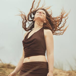 May include: A woman wearing a brown halter top and a matching wrap skirt. Her long, reddish-brown hair is blowing in the wind. The outfit is simple and earthy, with a natural, bohemian vibe. The background is a sandy beach.