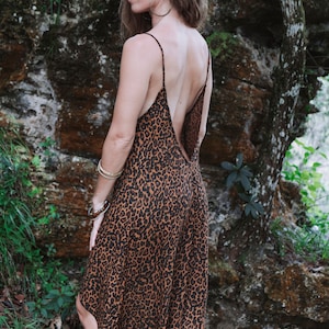Modèle portant une robe à bretelles mi-longue imprimé léopard marron, vue de dos, dos-nu plongeant et longueur asymétrique. Décor nature en forêt, plantes et roches humides.