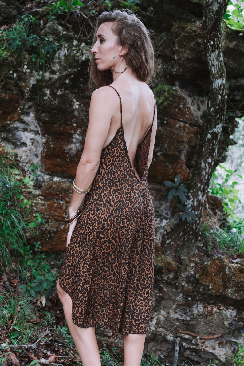Modèle portant une robe à bretelles mi-longue imprimé léopard marron, vue de dos, dos-nu plongeant et longueur asymétrique. Décor nature en forêt, plantes et roches humides.