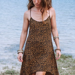 Modèle portant une robe à bretelles mi-longue, coupe asymétrique, imprimé léopard marron, vue de face. Décor bord de lac, herbe et sable, arbres au loin.