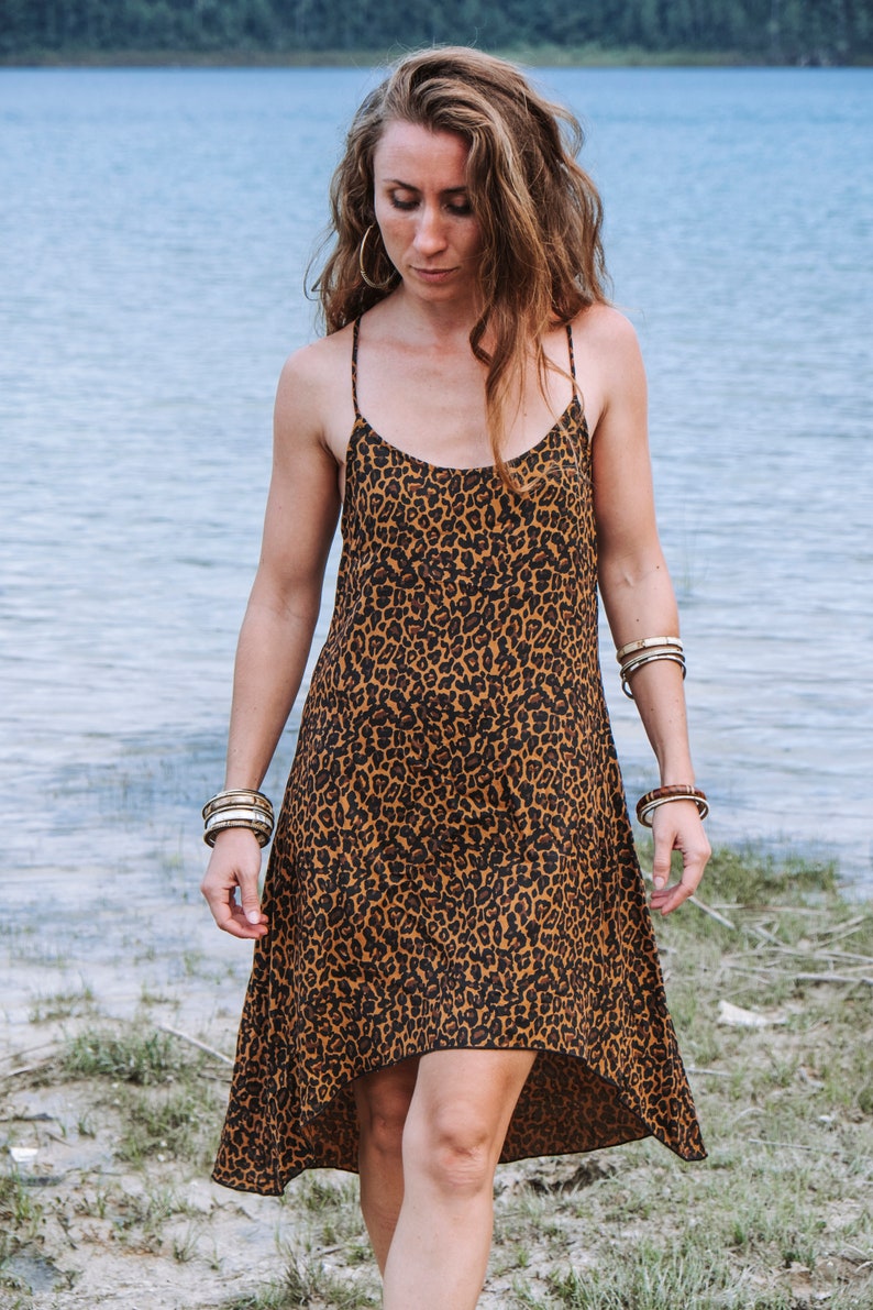 Modèle portant une robe à bretelles mi-longue, coupe asymétrique, imprimé léopard marron, vue de face. Décor bord de lac, herbe et sable, arbres au loin.
