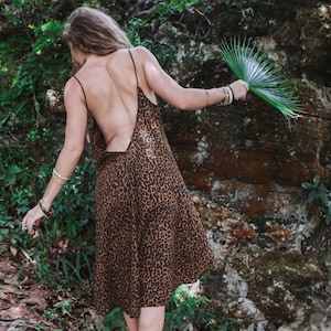 Modèle en mouvement portant une robe à bretelles mi-longue imprimé léopard marron, vue de dos, dos-nu plongeant en V. Décor nature en foret, plantes exotiques et roches humides