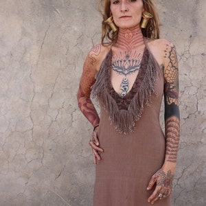 Könnte beinhalten: Eine Frau in einem braunen Kleid mit kunstvollen Tätowierungen an Armen und Hals. Sie trägt große goldene Ohrringe und einen Nasenring.