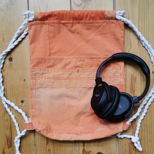 Op de afbeelding: Een oranje rugzak met witte touwriemen en een zwarte over-ear koptelefoon. De rugzak heeft een zakontwerp aan de voorkant.